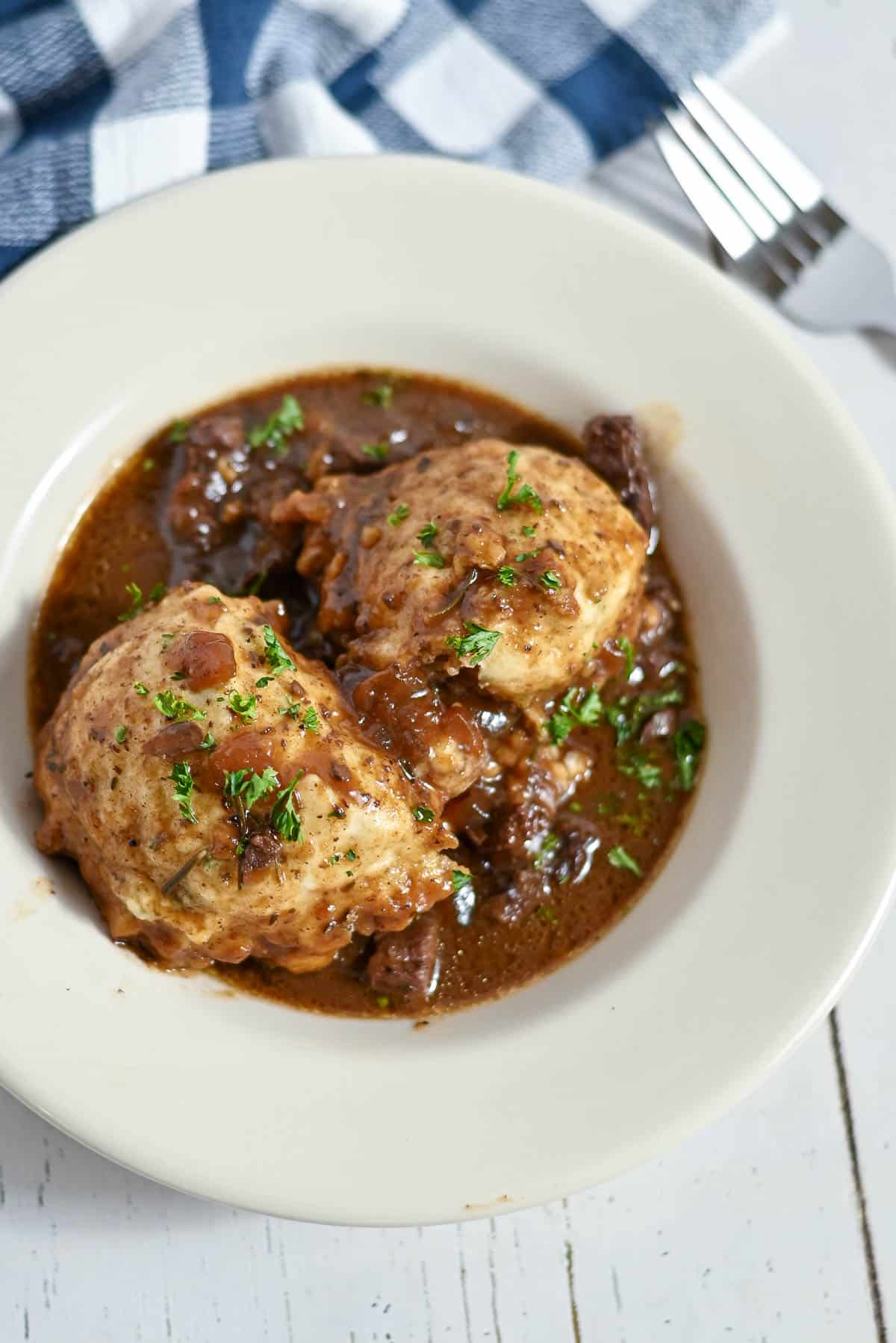 Two dumplings in rich brown stew, garnished with herbs, served on a white plate with a fork and napkin nearby.