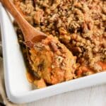 Sweet potato casserole topped with a pecan crumble, served in a white baking dish with a wooden spoon.