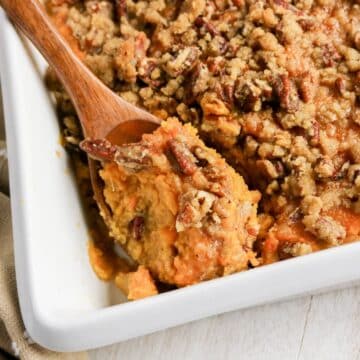 Sweet potato casserole topped with a pecan crumble, served in a white baking dish with a wooden spoon.