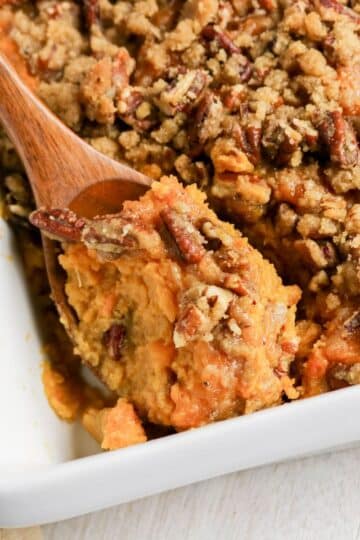 Sweet potato casserole topped with a pecan crumble, served in a white baking dish with a wooden spoon.