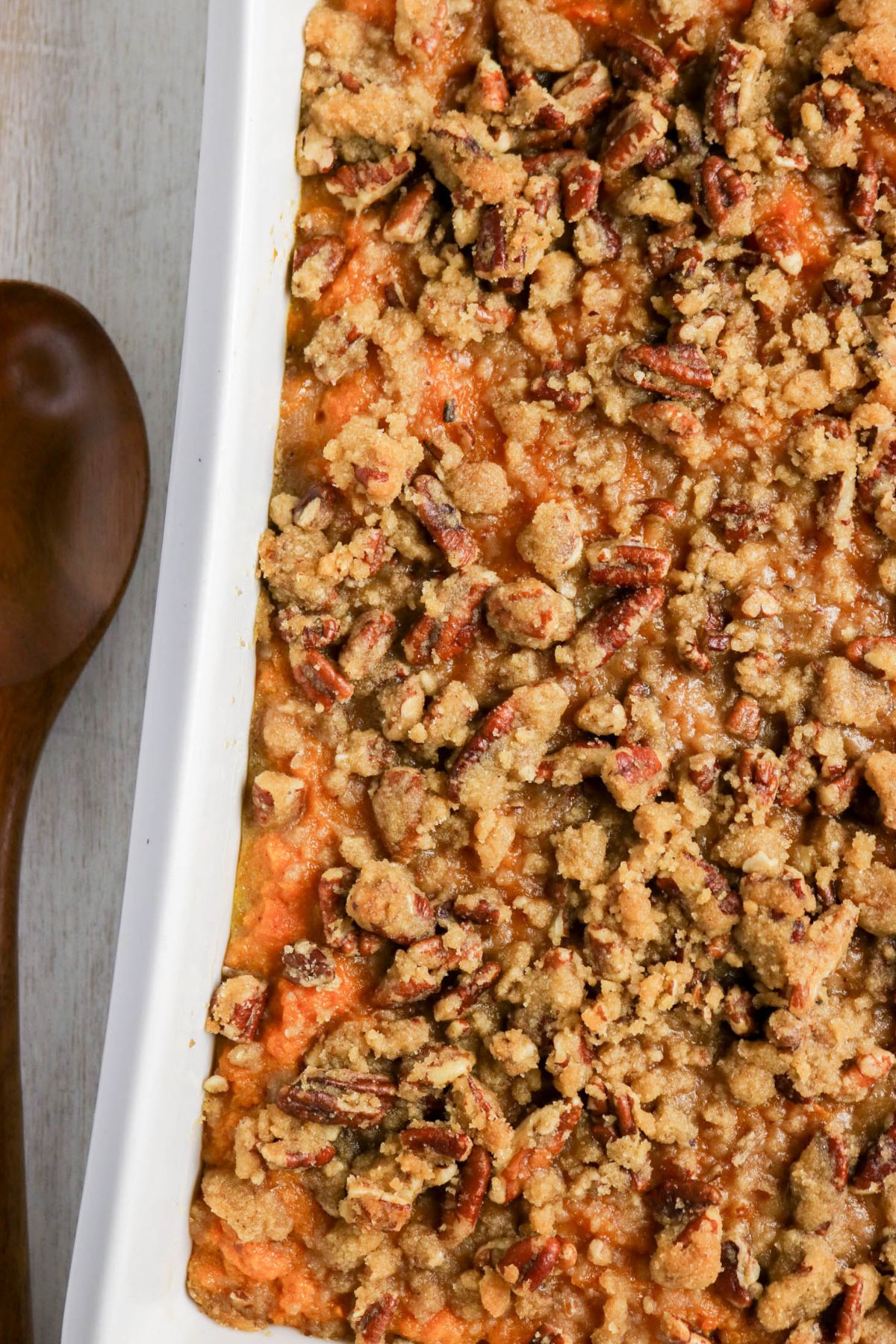Sweet potato casserole topped with a crumbly pecan streusel in a white baking dish beside a wooden spoon.