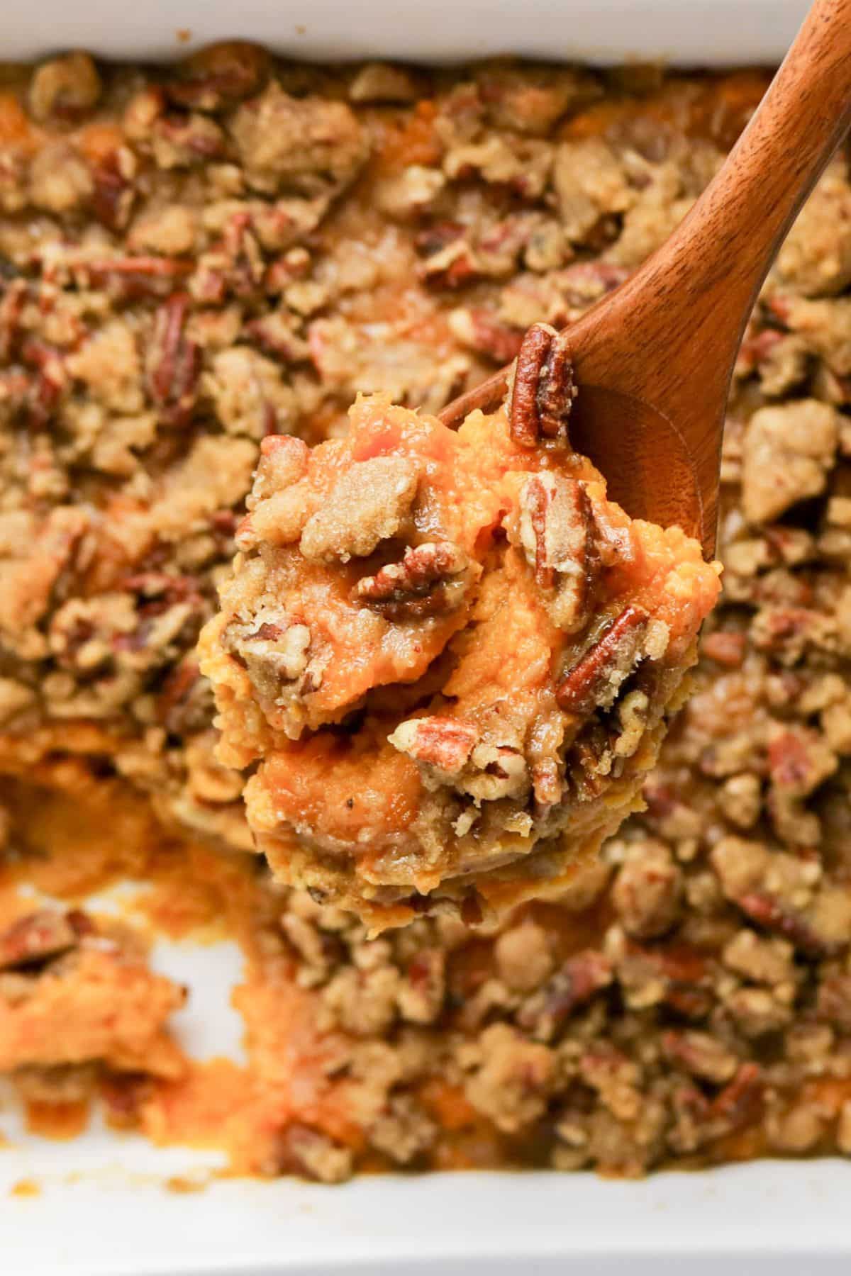 A wooden spoon holds a serving of sweet potato casserole topped with a brown sugar pecan crumble.