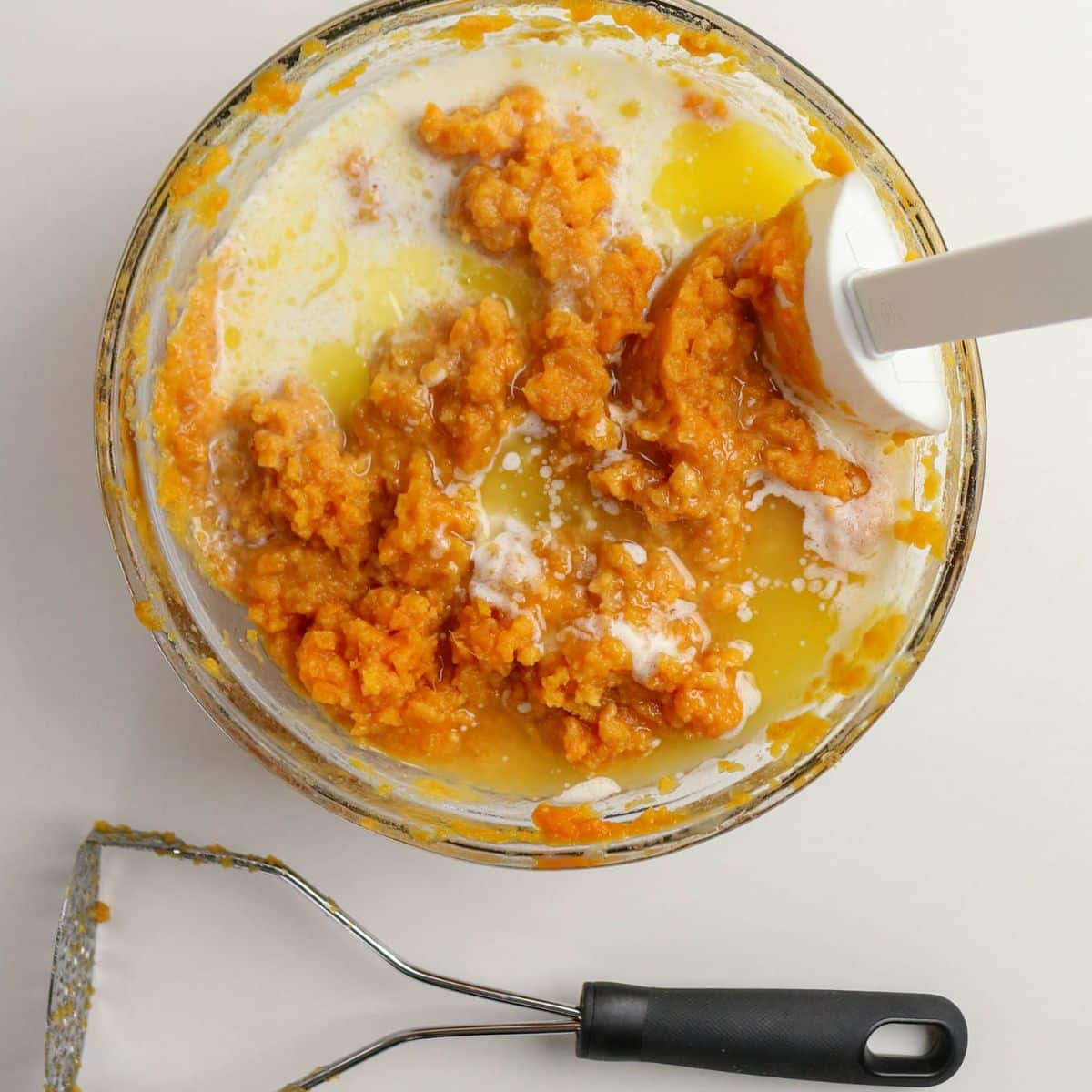Bowl of mashed sweet potatoes with melted butter and milk, spatula inside, and a potato masher nearby.