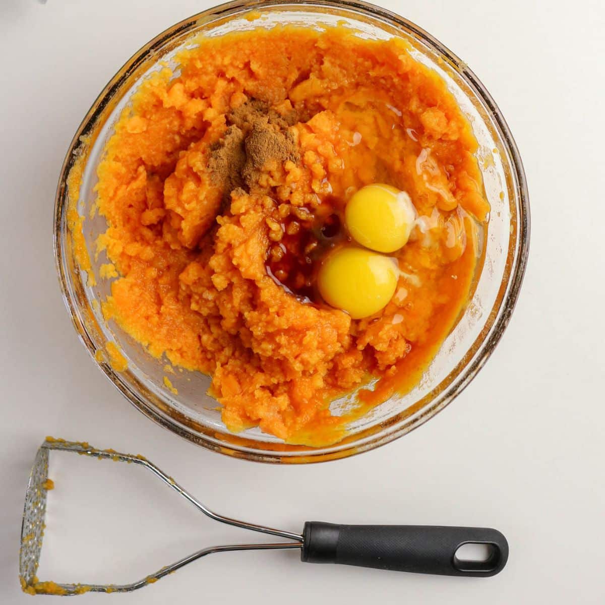 A bowl with mashed sweet potatoes, two eggs, cinnamon, and vanilla, next to a potato masher on a white surface.
