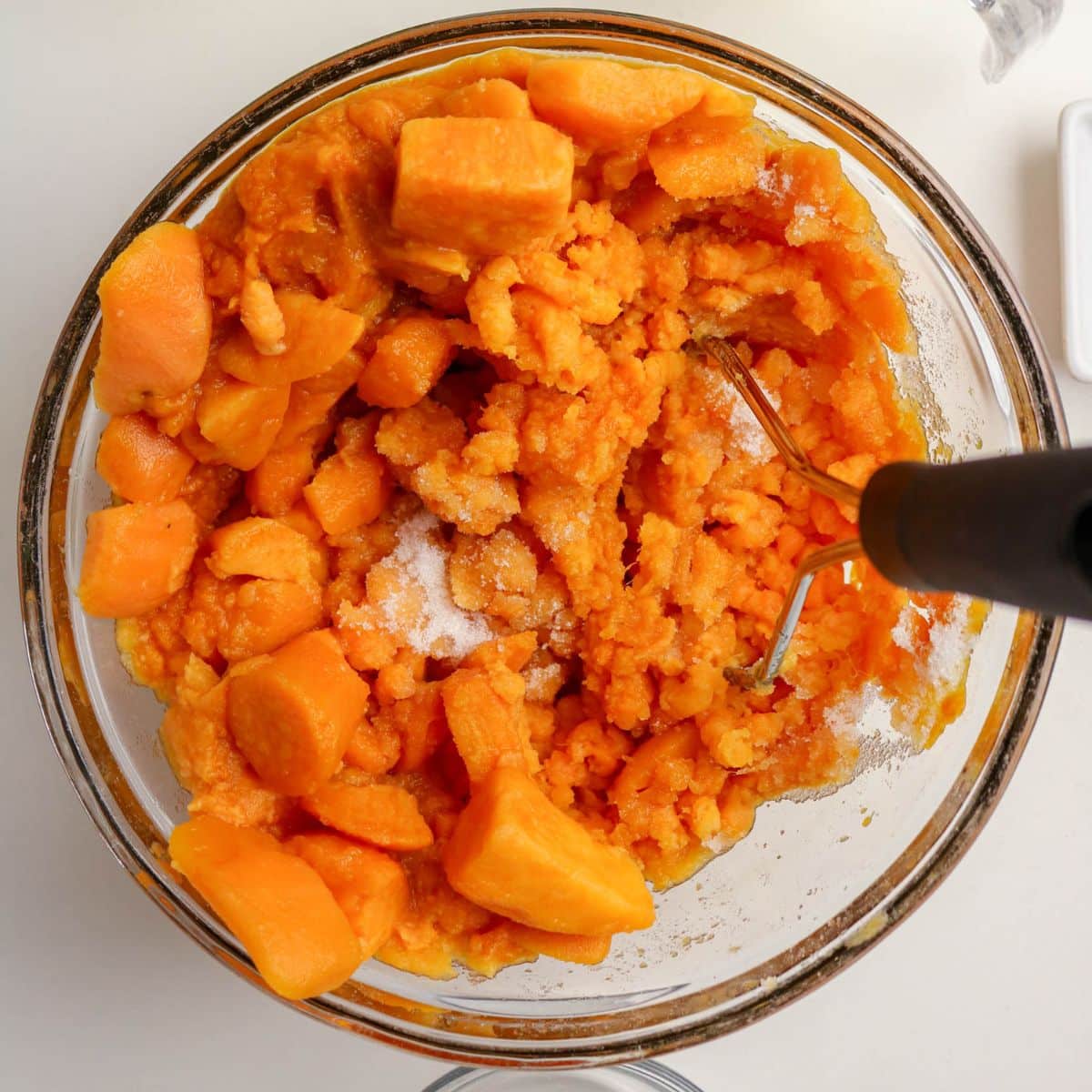 A bowl of cubed, mashed sweet potatoes with a masher and a sprinkle of sugar on top.