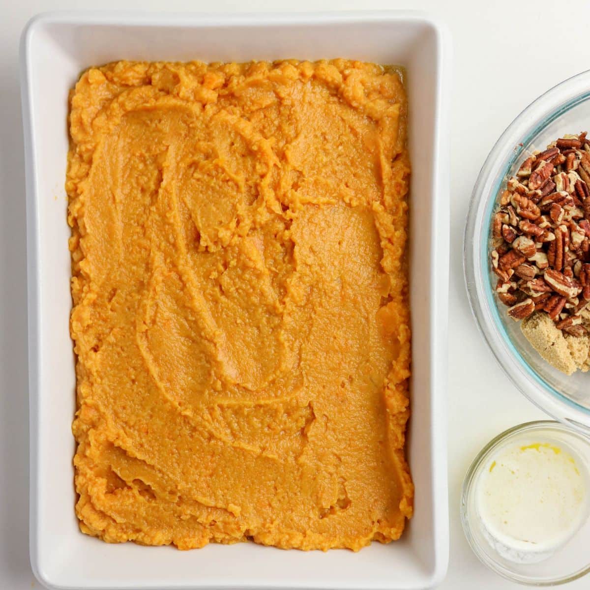 Mashed sweet potatoes spread in a white baking dish, with pecans and brown sugar in bowls nearby.