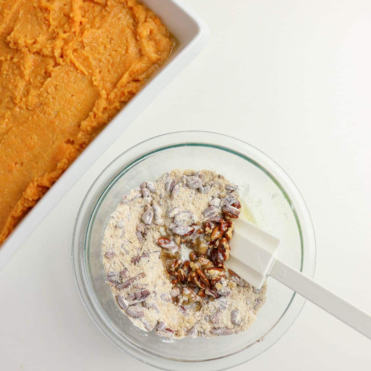 A bowl of flour, pecans, and chocolate chips beside a dish of orange sweet potato mixture on a white surface.