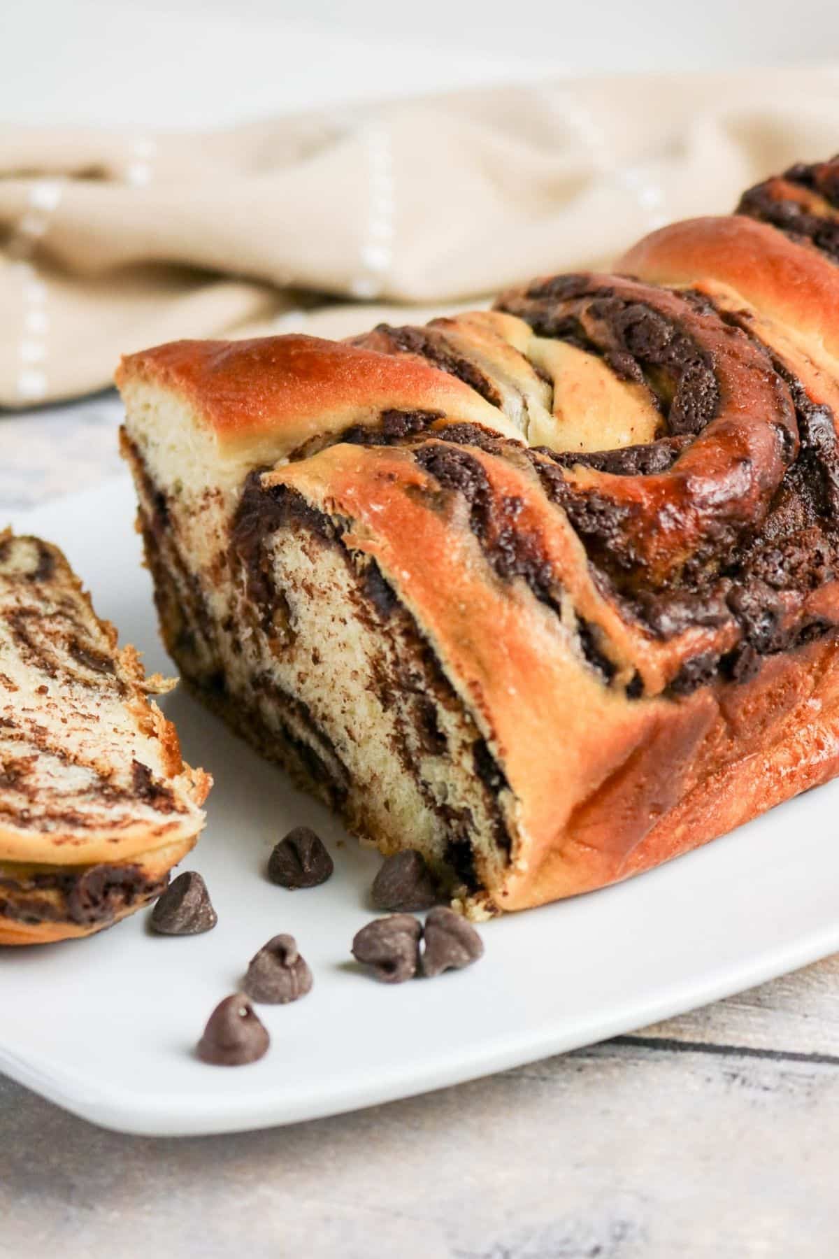 A sliced chocolate swirl bread loaf sits on a white plate, with chocolate chips beside it.