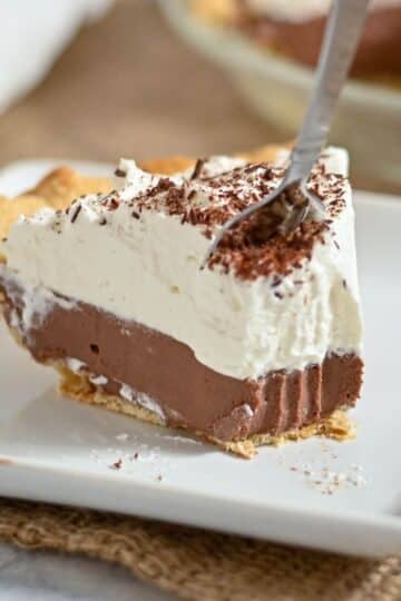 A fork cutting into a slice of chocolate cream pie with whipped topping on a white plate.