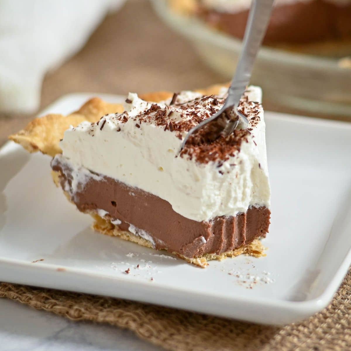 A fork cutting into a slice of chocolate cream pie with whipped topping on a white plate.