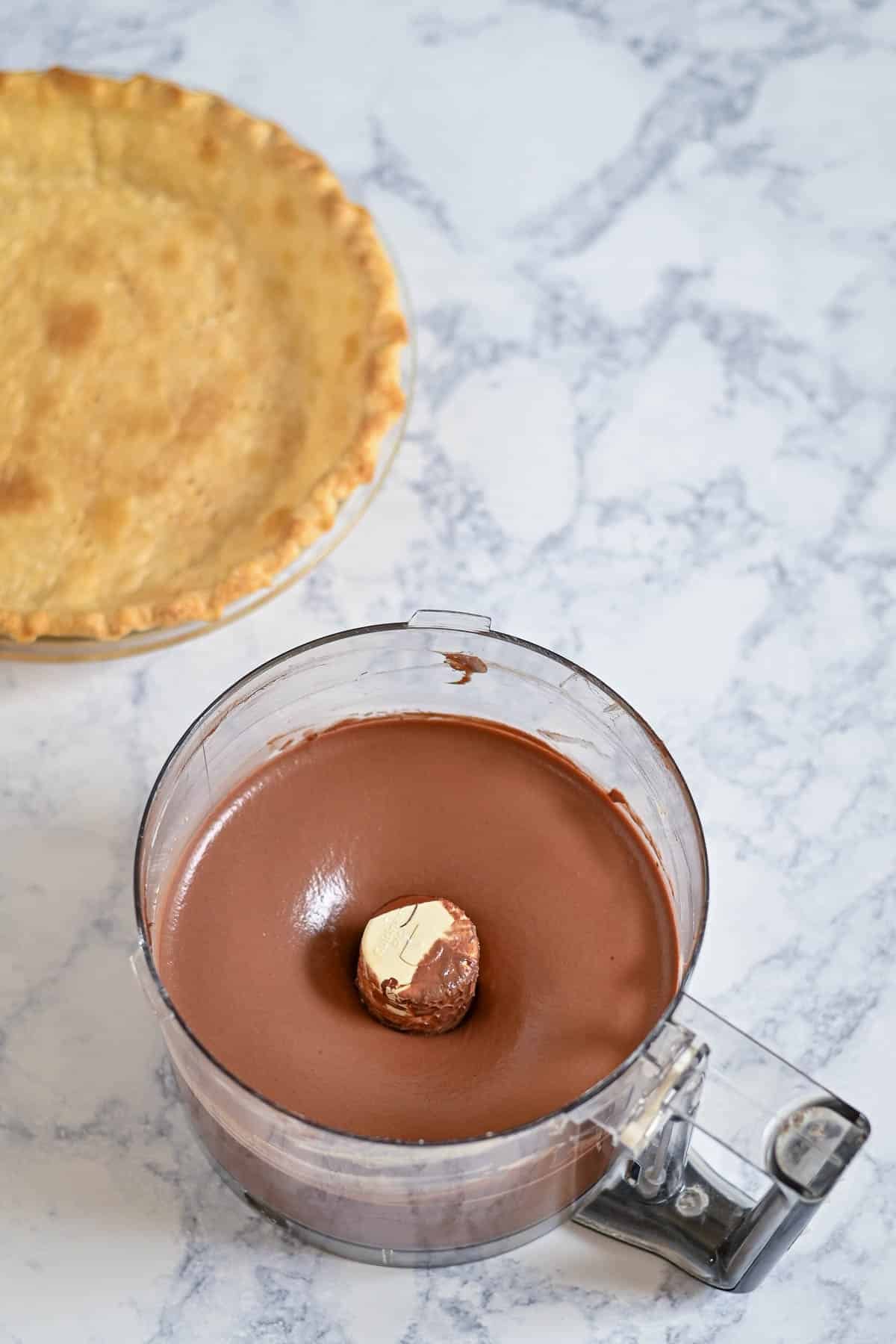 A food processor with chocolate filling and butter next to an empty baked pie crust on a marble surface.