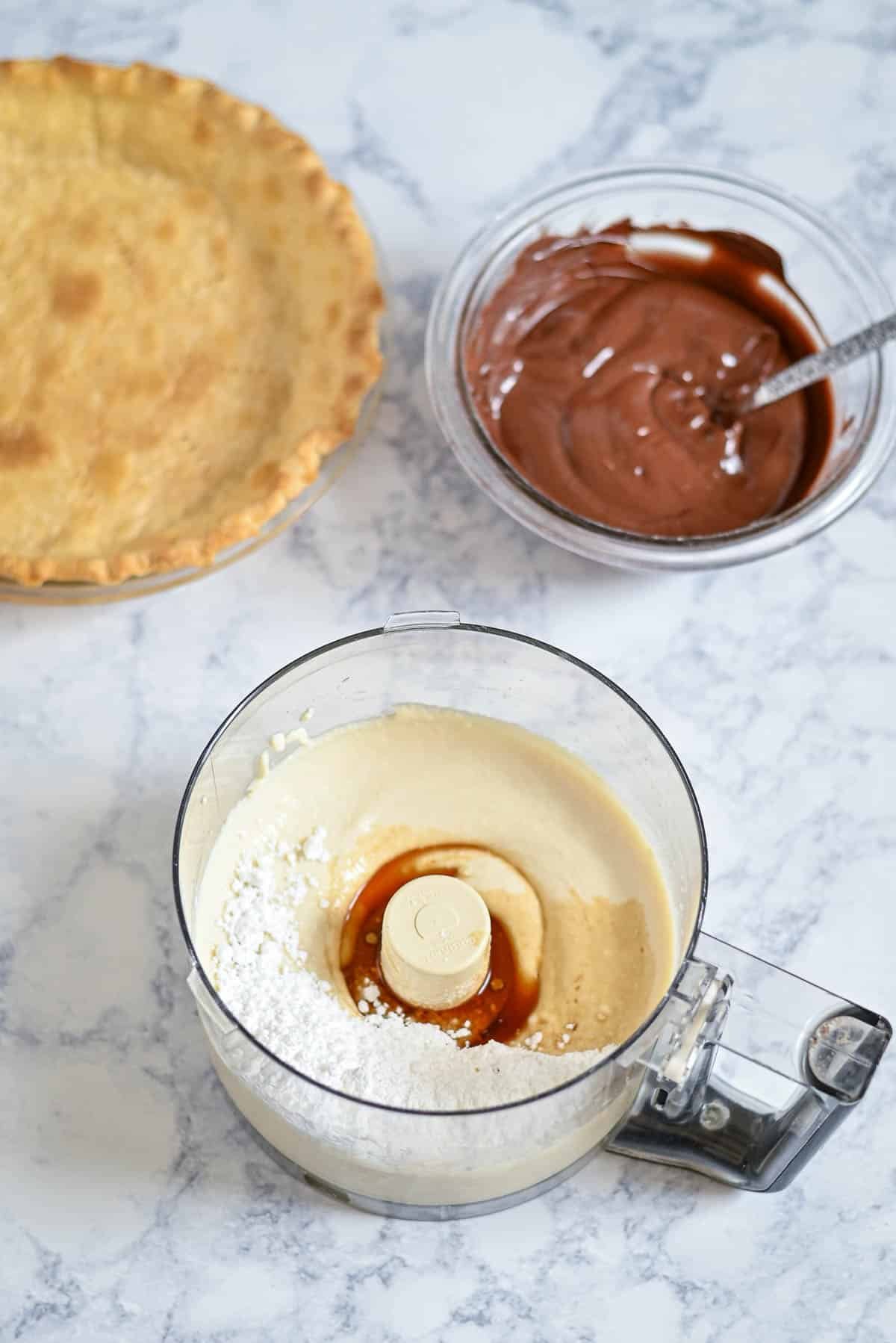 A pie crust, chocolate mixture in a bowl, and creamy filling in a food processor on a marble counter.