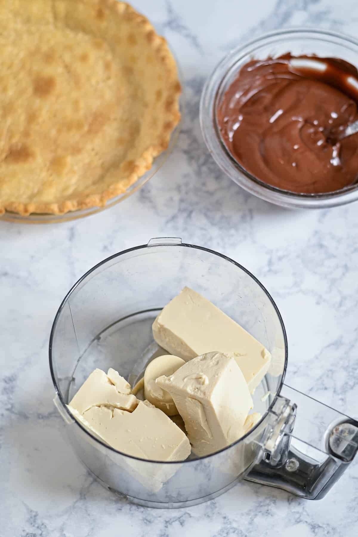 Pie crust, melted chocolate in a bowl, and cream cheese in a food processor on a marble surface.
