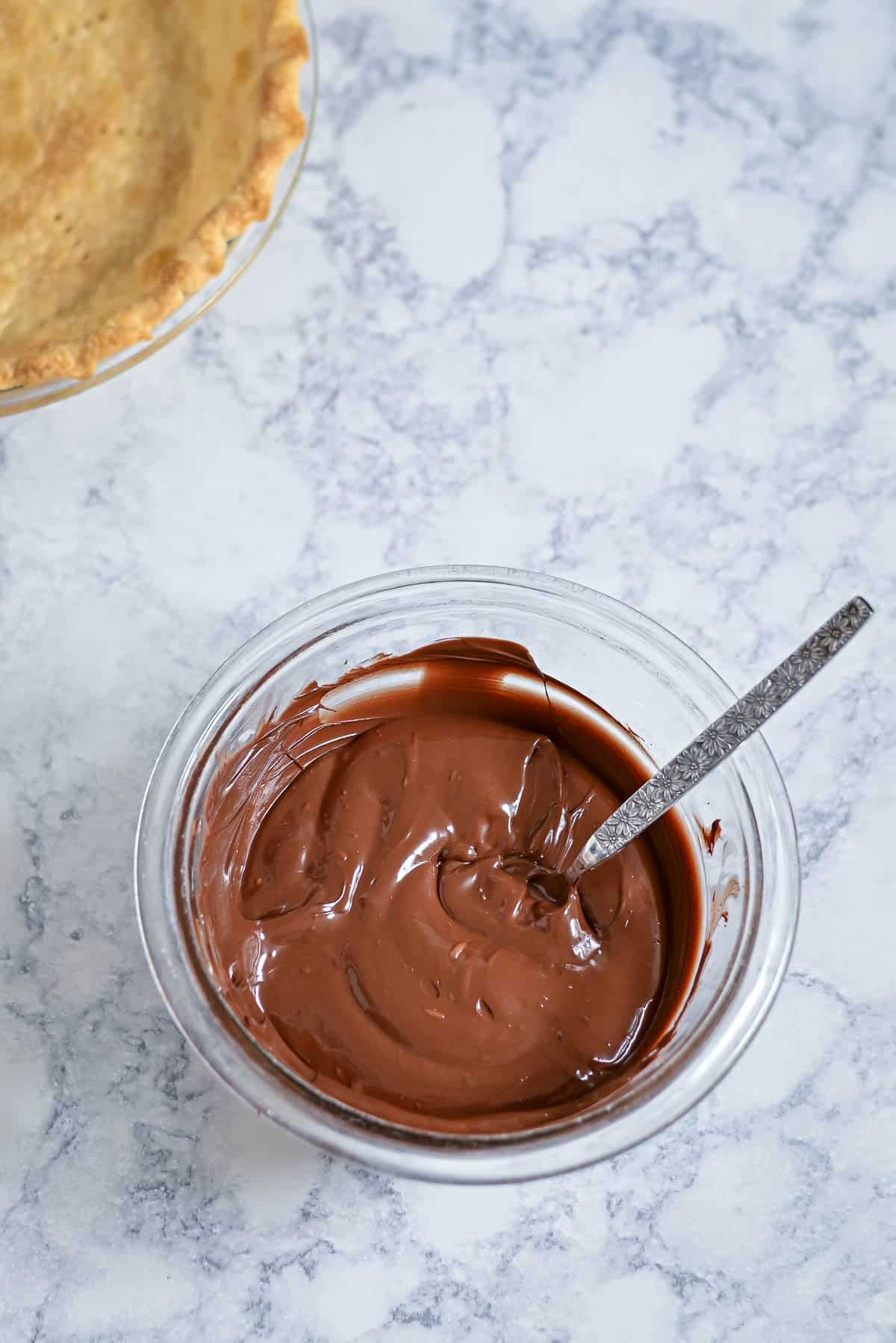 A glass bowl of melted chocolate with a spoon, next to a partially visible baked pie crust.