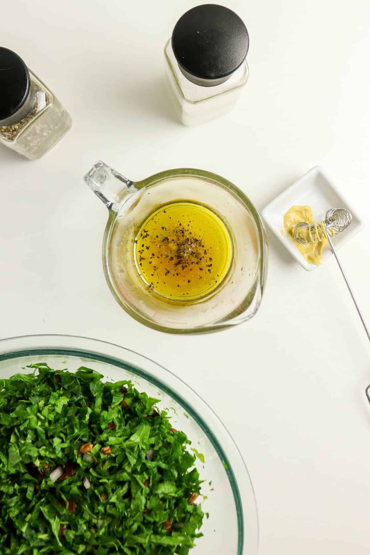 A bowl of chopped greens, a measuring cup with dressing, mustard, and salt and pepper shakers on a white surface.