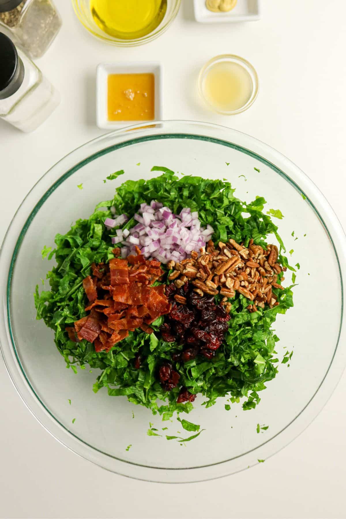 Chopped collard greens, bacon, red onion, pecans, and dried cranberries in a glass bowl, with dressing ingredients nearby.
