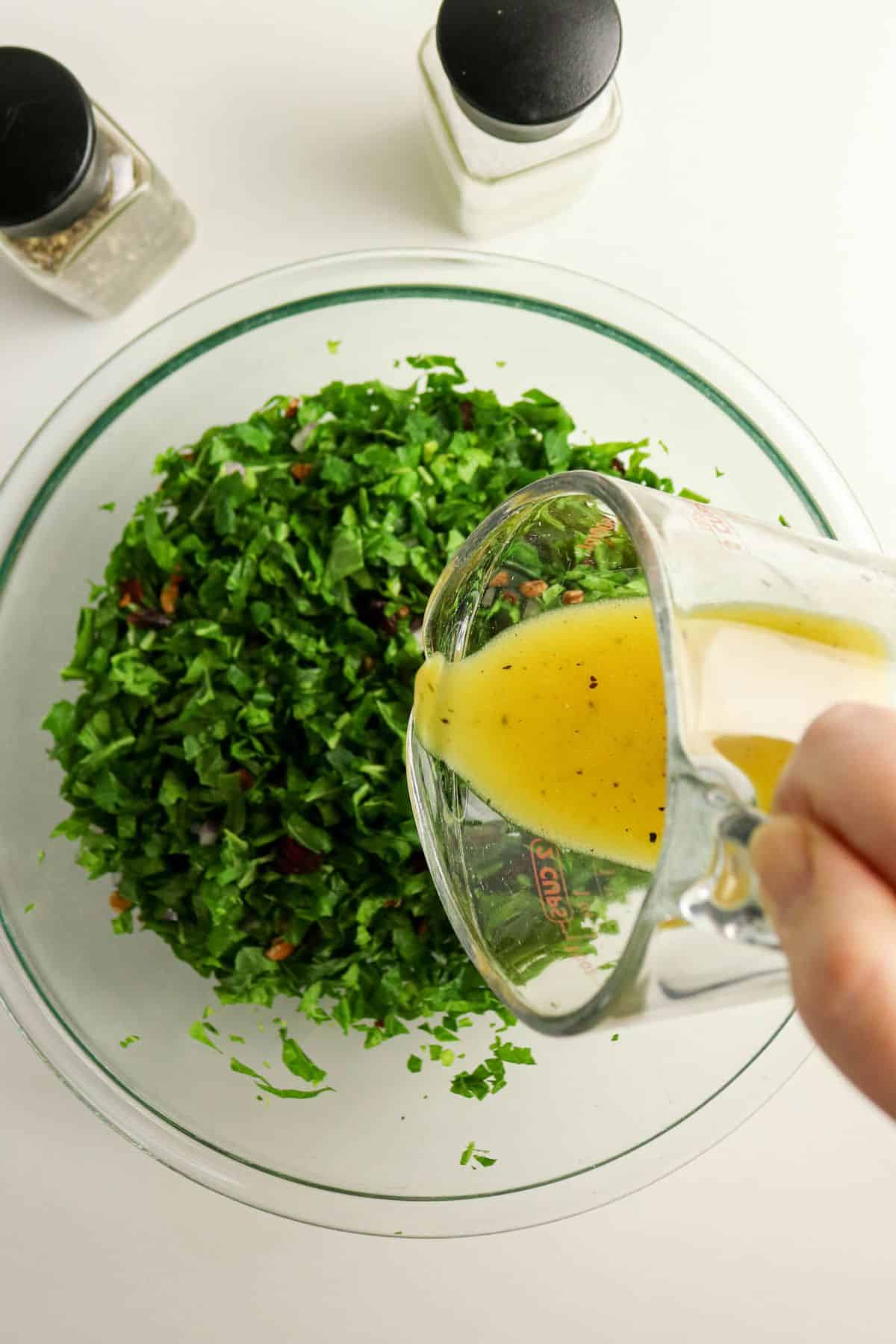 Hand pouring yellow dressing onto chopped greens in a glass bowl, with salt and pepper shakers nearby.