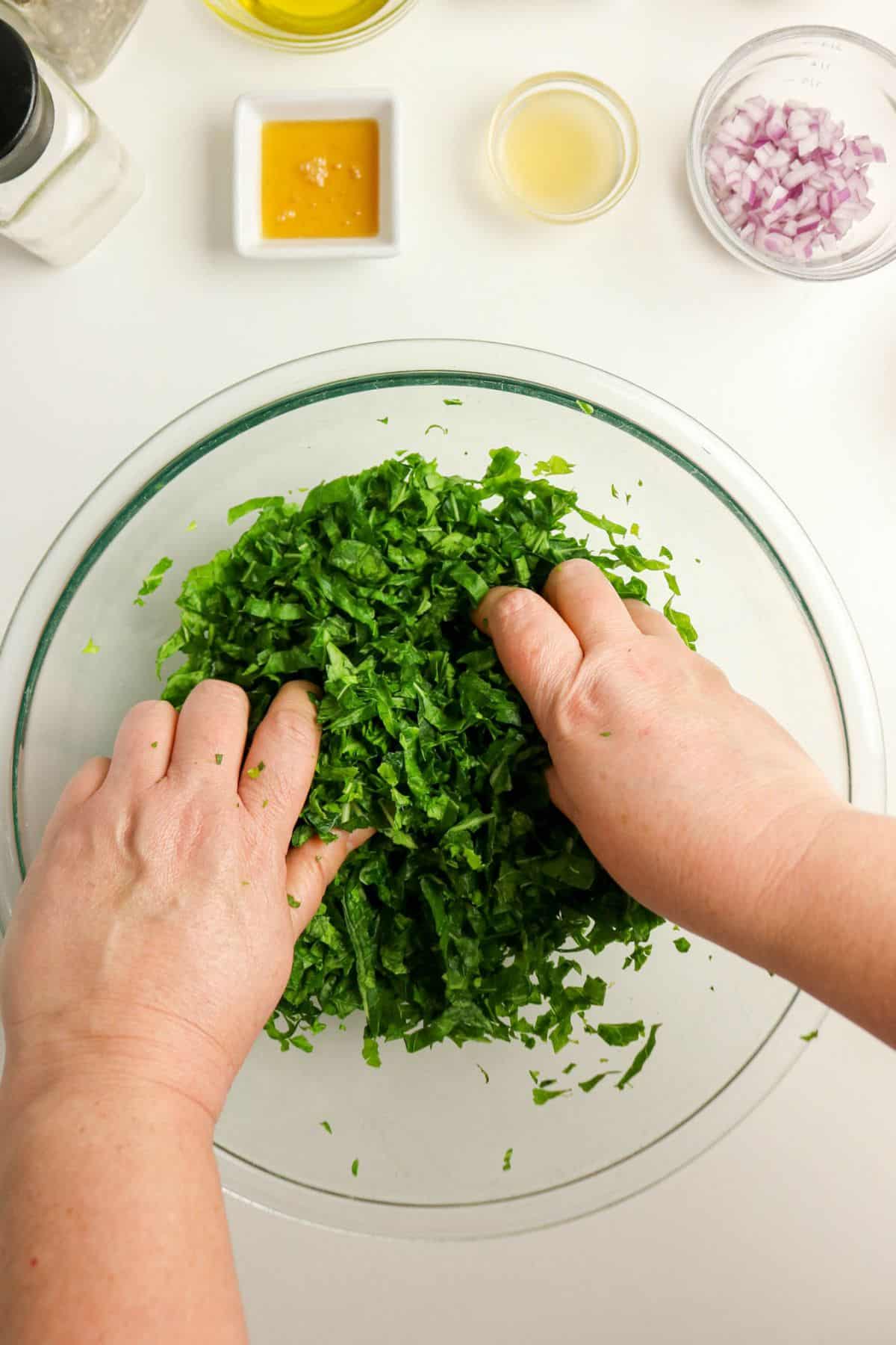 Hands massaging chopped collard greens in a glass bowl with salad ingredients on a white surface nearby.