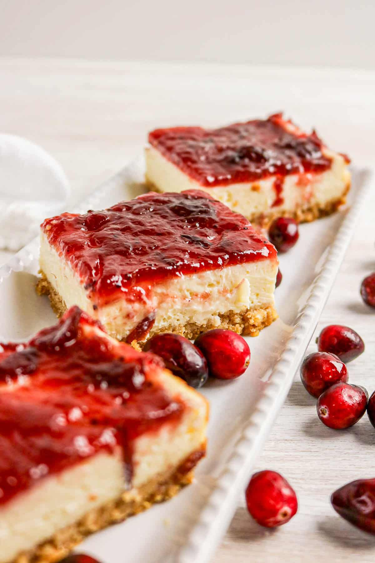 Three cheesecake bars topped with glossy red berry sauce, garnished with fresh cranberries on a white platter.