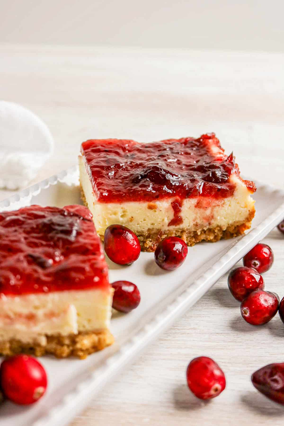 Two cheesecake bars topped with cranberry sauce on a white plate, surrounded by fresh cranberries.