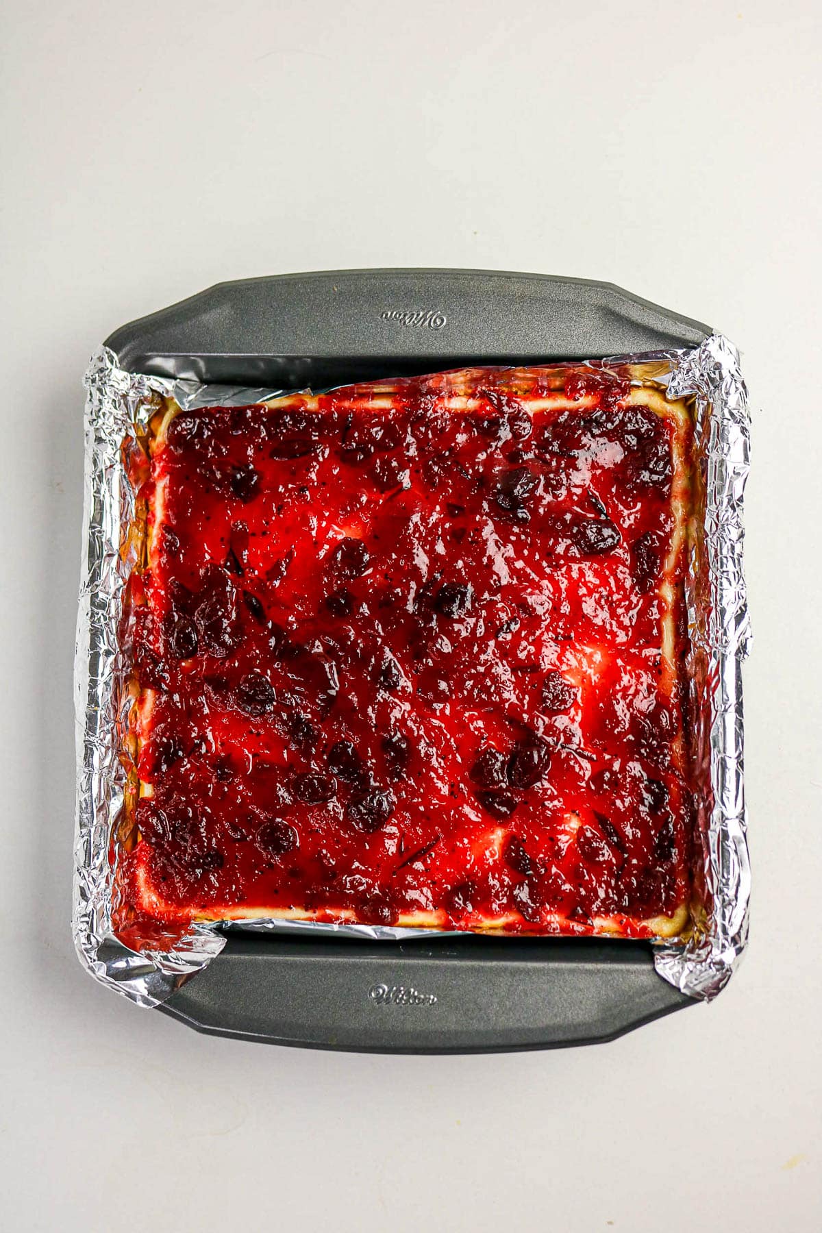 A square baking pan with a foil lining holds a dessert topped with a glossy red fruit glaze.