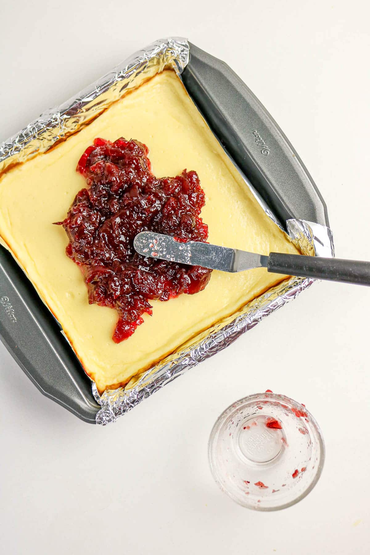 A spatula spreads jam over cheesecake in a foil-lined baking pan; an empty jar sits nearby.