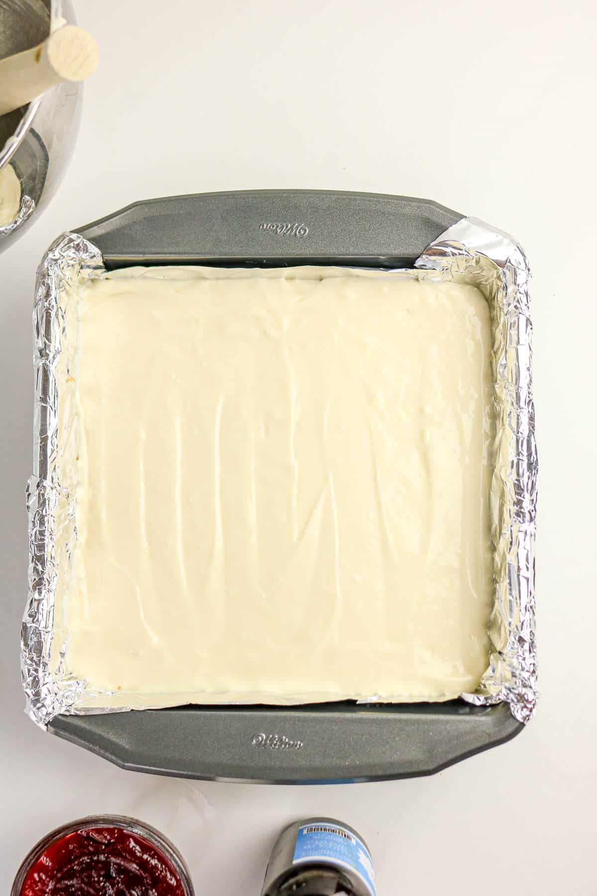 Square baking pan lined with foil, filled with smooth cream cheese mixture; jelly jar and bottle nearby.