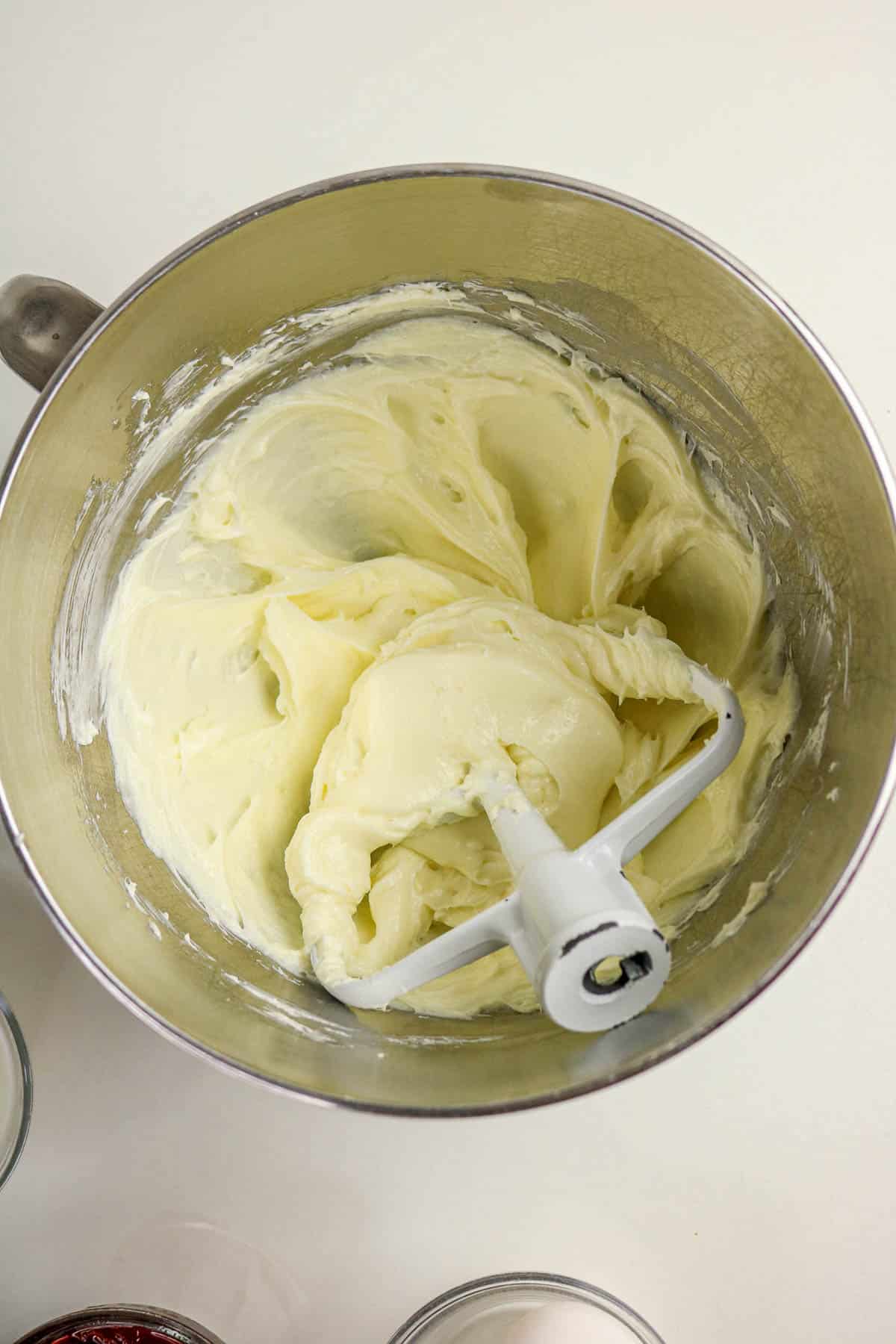 Mixing bowl with creamy white frosting and a paddle attachment on a white surface.