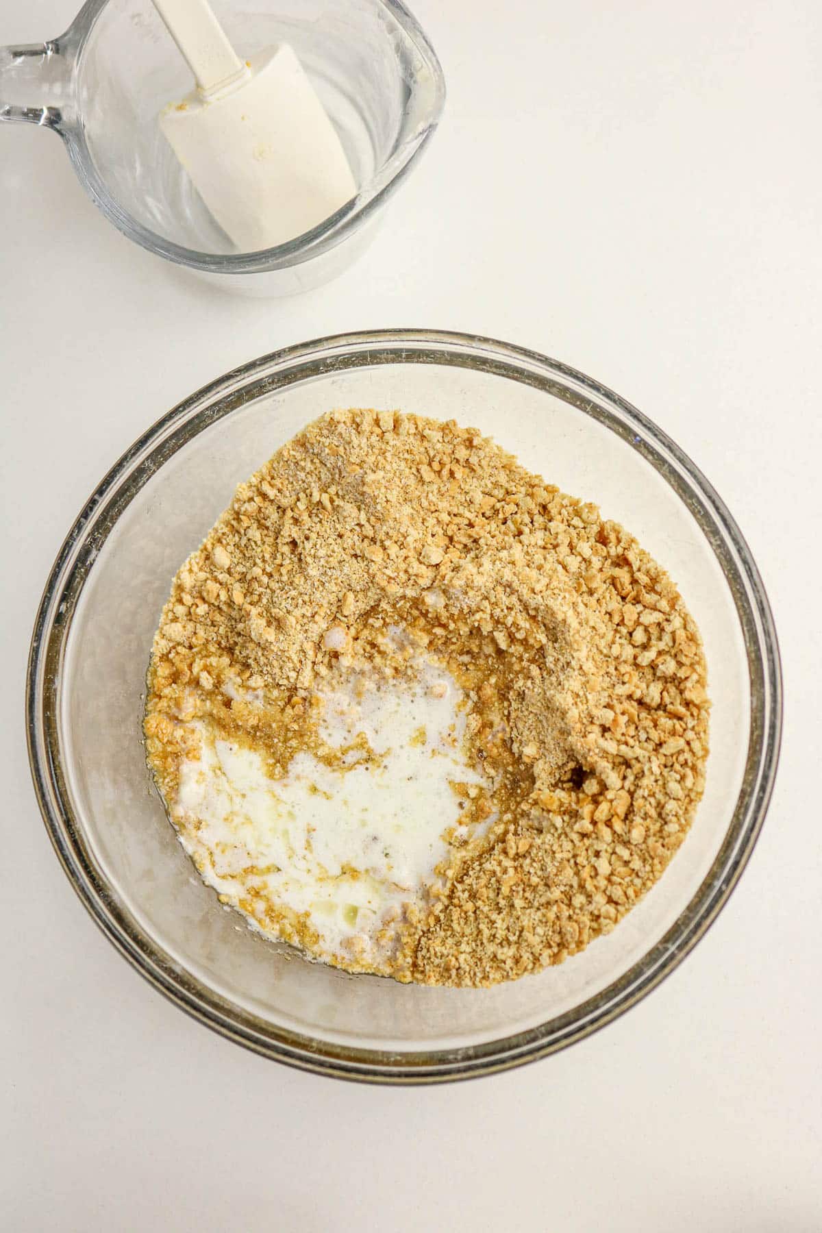 A glass bowl with graham cracker crumbs, melted butter, and milk next to an empty bowl with a spatula.