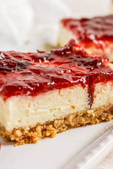 A slice of cheesecake with a crumbly crust and a glossy red berry topping on a white plate.