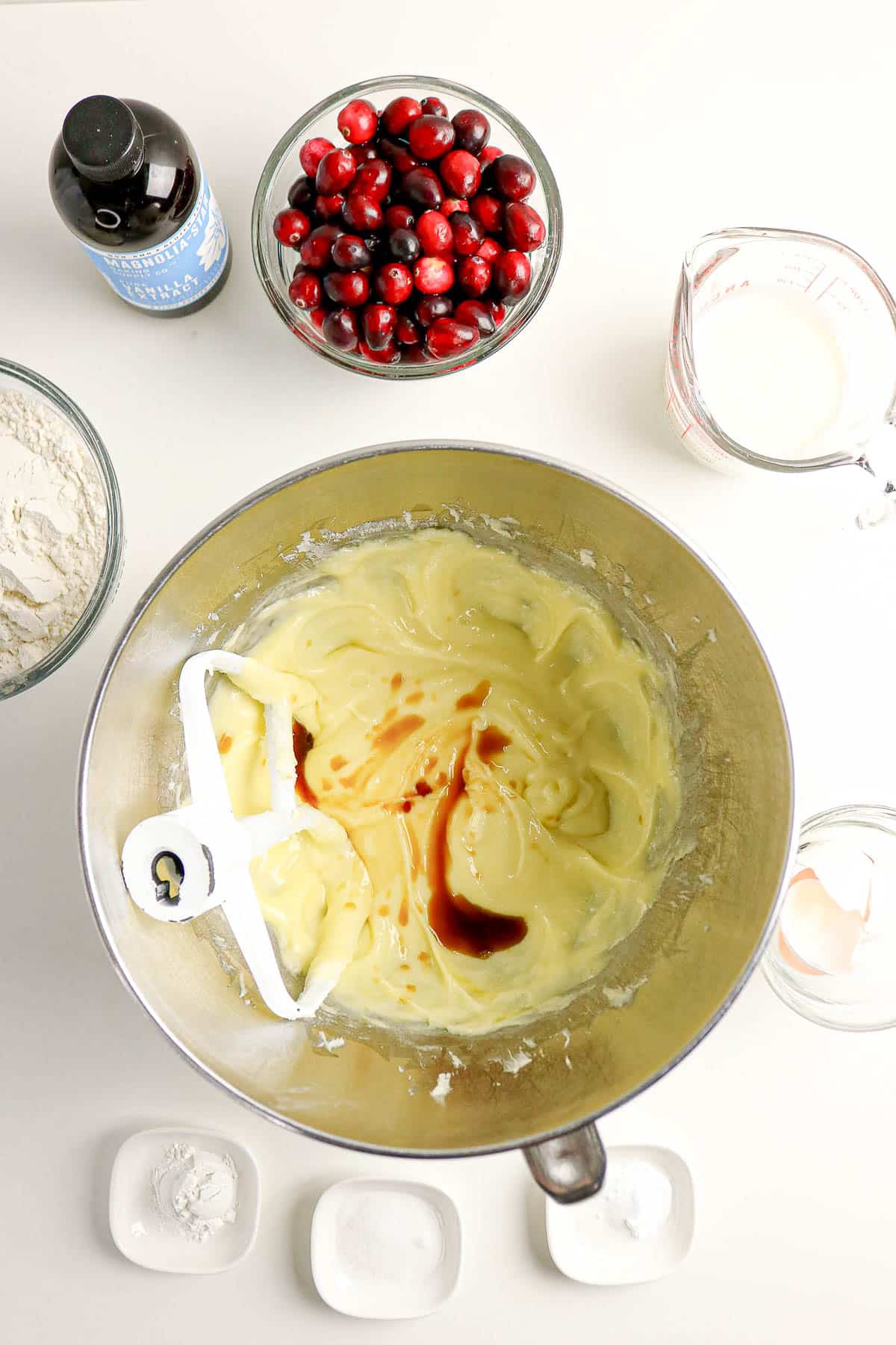 Mixing bowl with batter and vanilla, surrounded by cranberries, flour, milk, eggs, and baking ingredients on a counter.