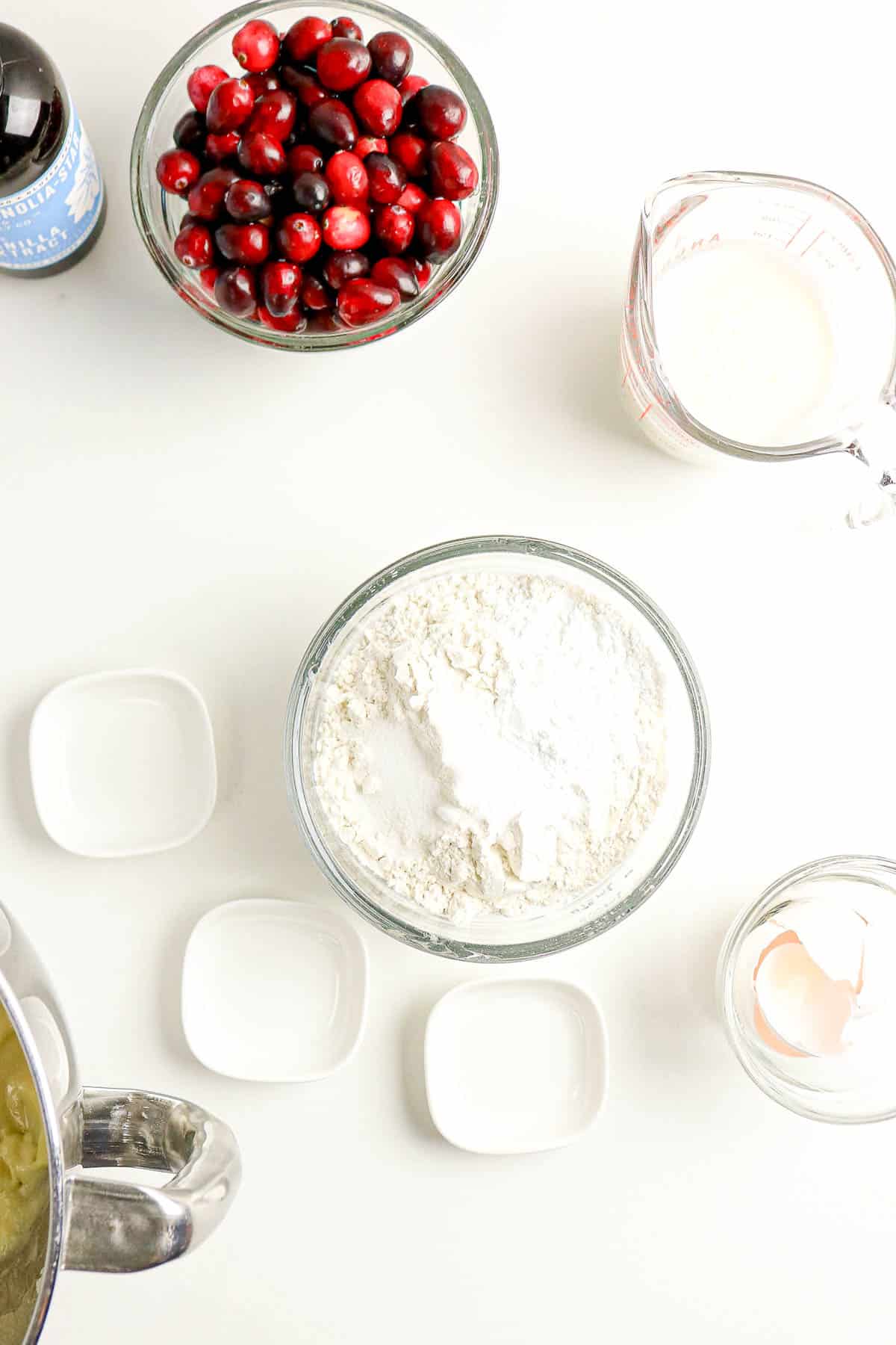 Bowl of cranberries, flour, measuring cup of milk, egg shell, and vanilla on a white surface.
