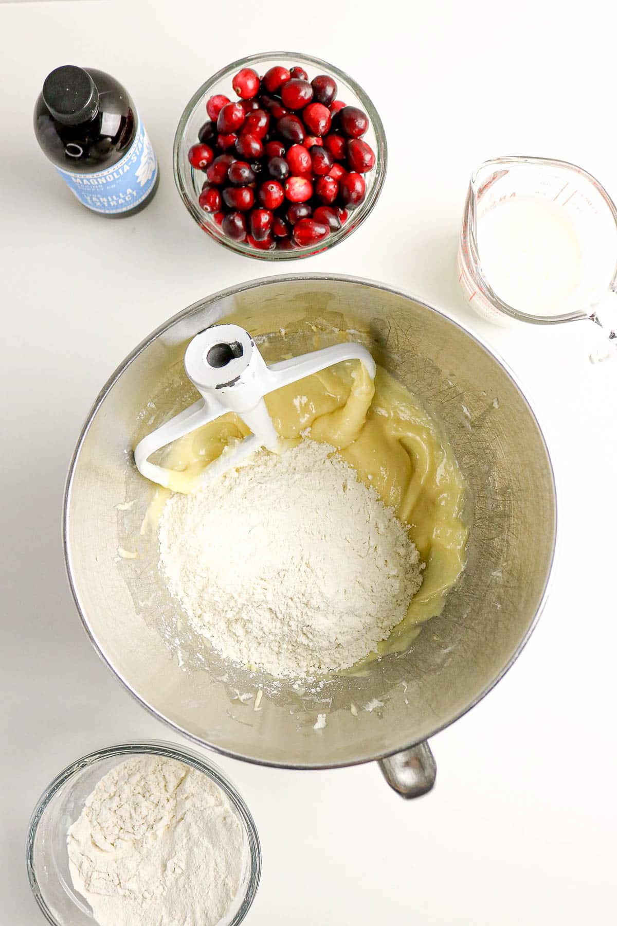 A mixing bowl with batter and flour, surrounded by cranberries, flour, vanilla, and milk on a white surface.