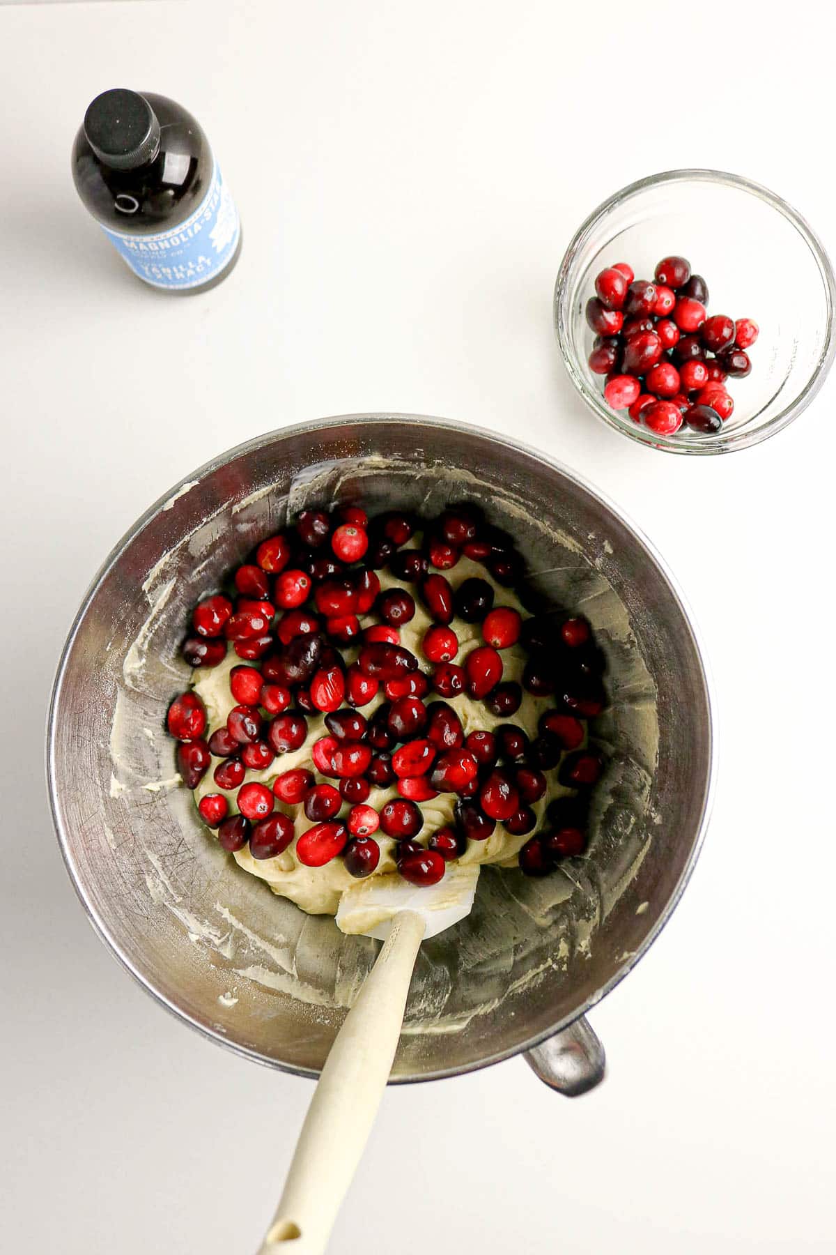 A mixing bowl with batter and cranberries, a spatula, a bowl of cranberries, and a bottle of vanilla extract.