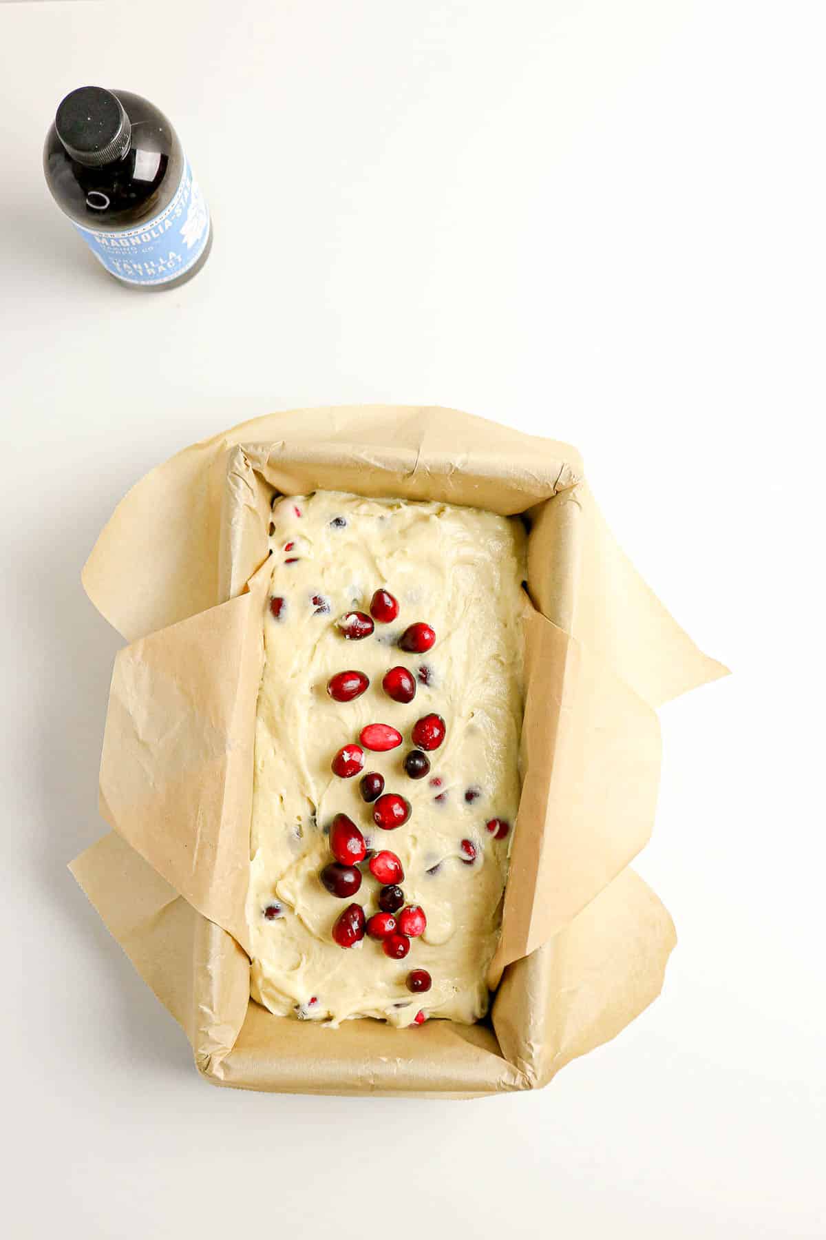 A loaf pan with unbaked batter topped with cranberries, next to a bottle of vanilla extract on a white surface.