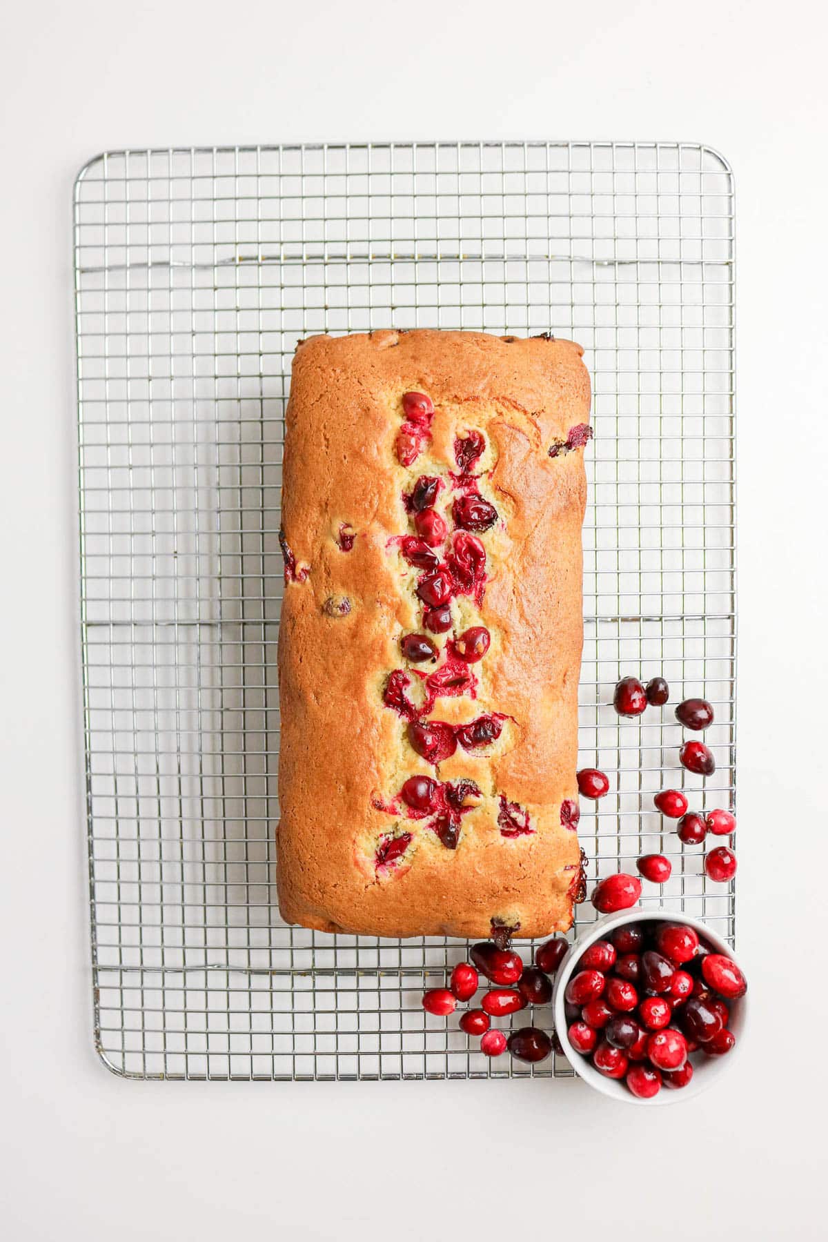 A loaf of cranberry bread on a cooling rack, with fresh cranberries scattered and in a small bowl nearby.