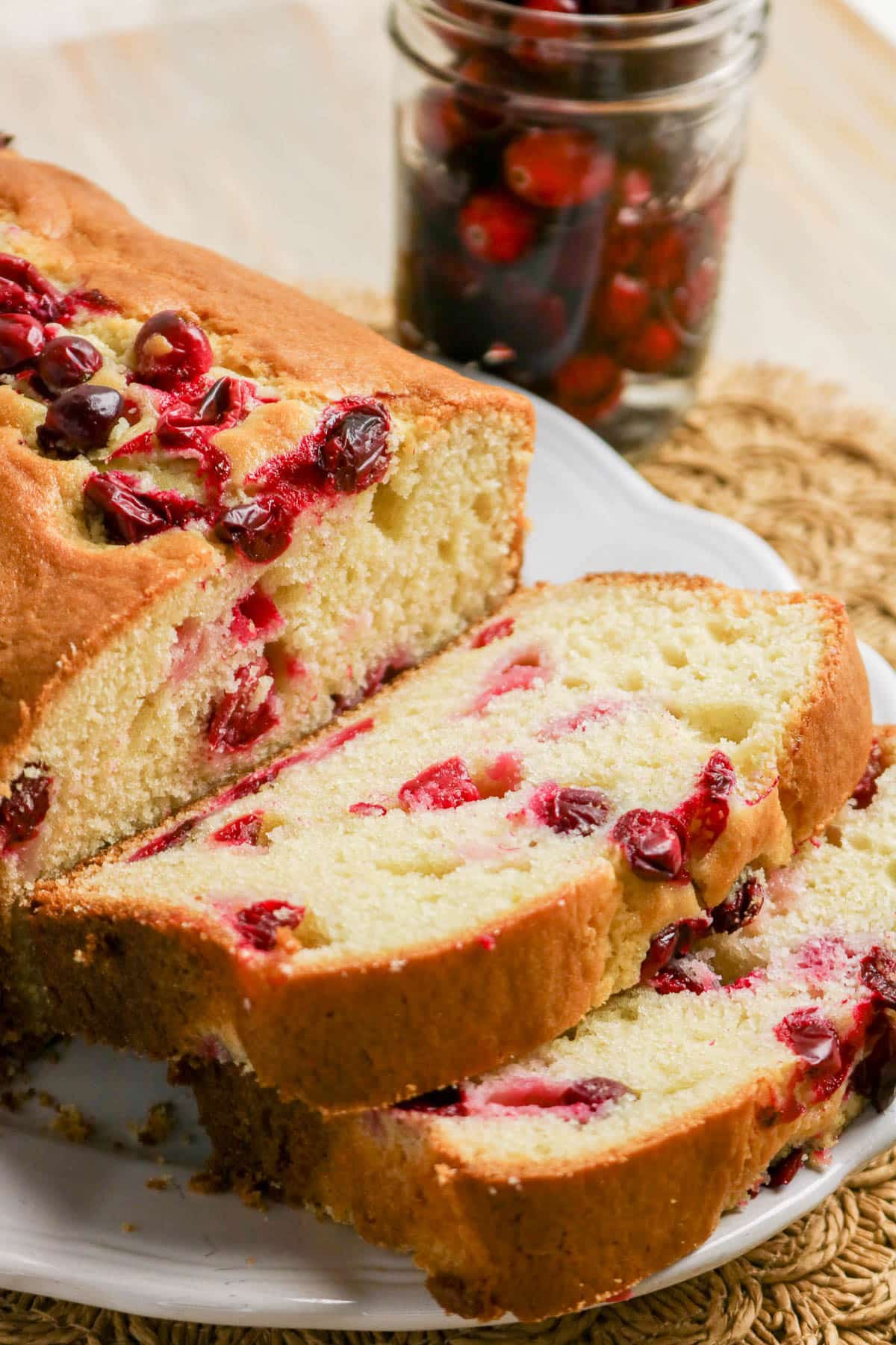 Sliced cranberry bread on a white plate with a jar of fresh cranberries in the background.