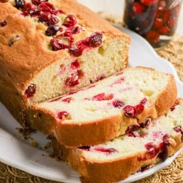 Sliced cranberry bread loaf on a white plate, showing cranberries inside and on top of the bread.