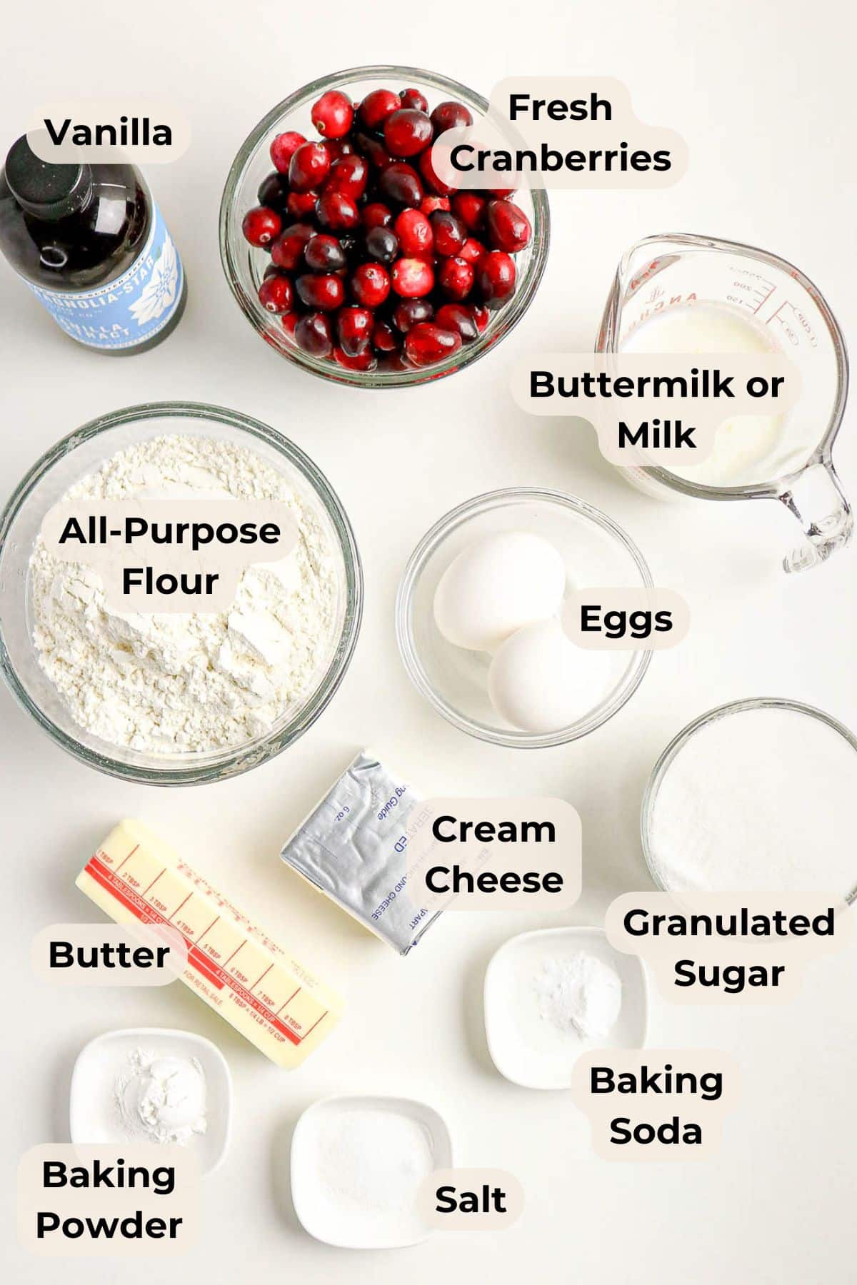 Baking ingredients labeled on a white surface, including cranberries, flour, eggs, butter, and cream cheese.