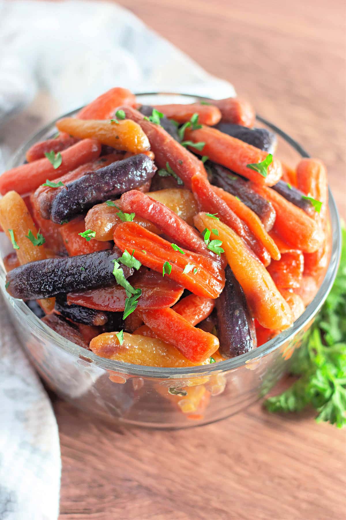 A glass bowl filled with glazed rainbow baby carrots, garnished with chopped parsley on a wooden surface.