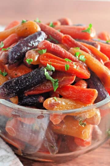 A glass bowl filled with colorful glazed baby carrots garnished with chopped parsley.