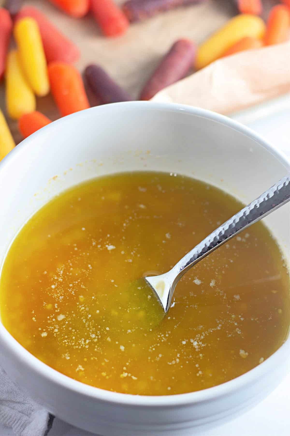 A bowl of yellow dipping sauce with a spoon, surrounded by colorful baby carrots in the background.