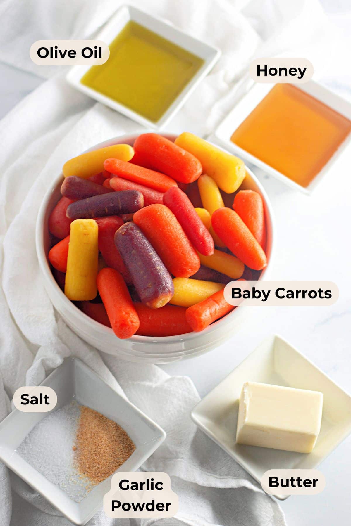 A bowl of colorful baby carrots surrounded by olive oil, honey, butter, salt, and garlic powder in small dishes.
