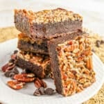 Three pecan-topped chocolate brownies stacked on a white plate with pecans and chocolate chips.