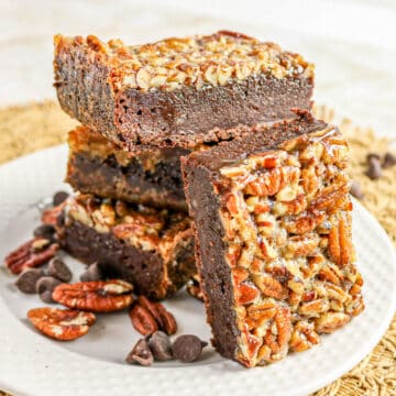 Three pecan-topped chocolate brownies stacked on a white plate with pecans and chocolate chips.