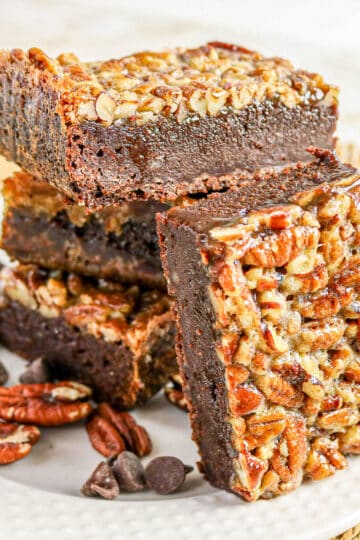 Three pecan-topped chocolate brownies stacked on a white plate with pecans and chocolate chips.