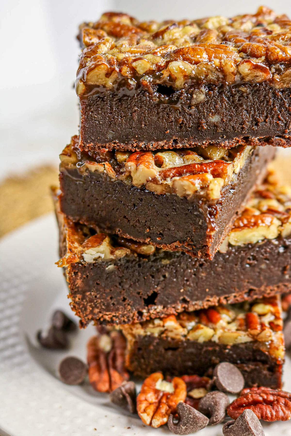 Three stacked pecan brownies with gooey topping, surrounded by chocolate chips and pecans on a white plate.