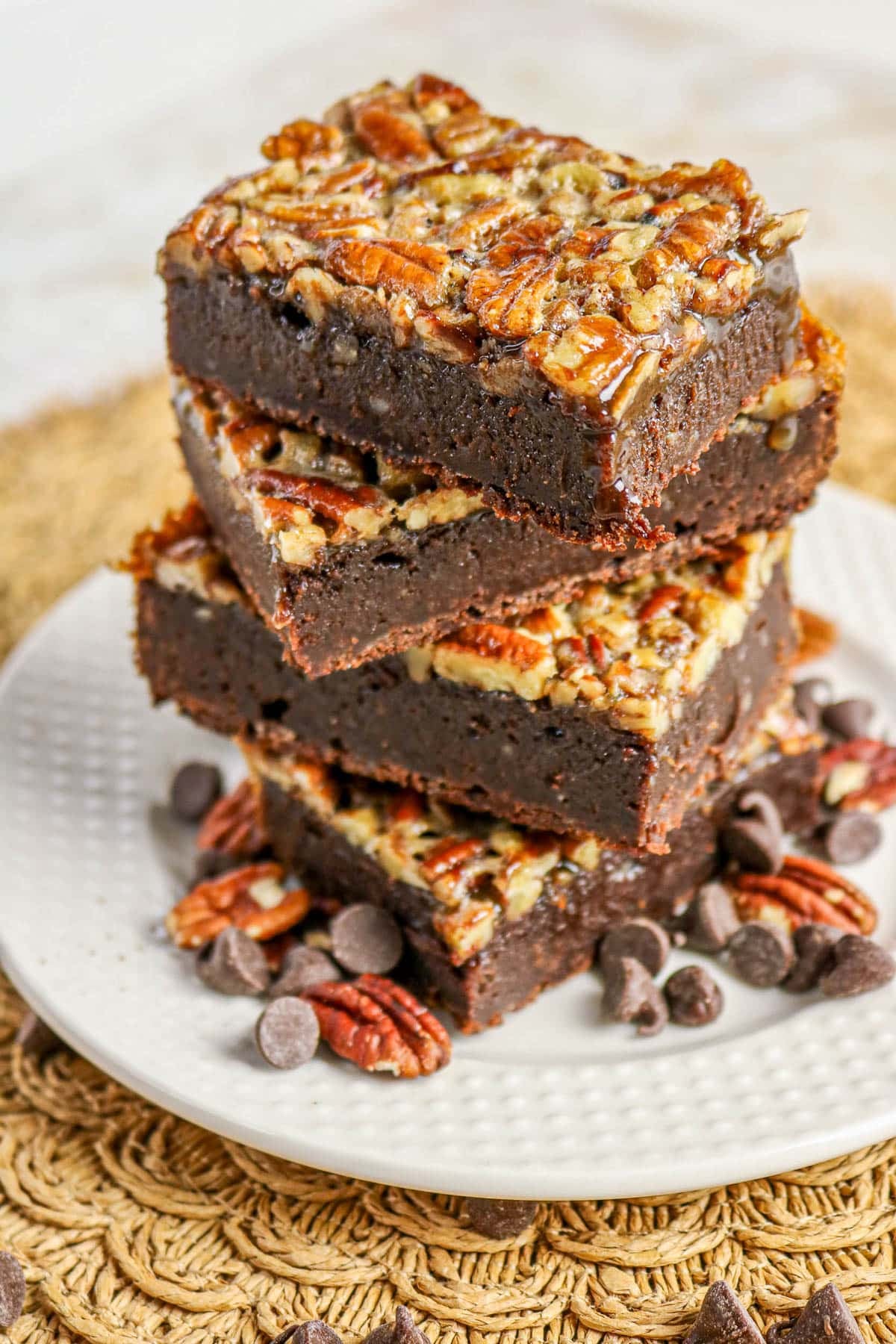 Three square pecan brownies stacked on a white plate with chocolate chips and pecans scattered around.