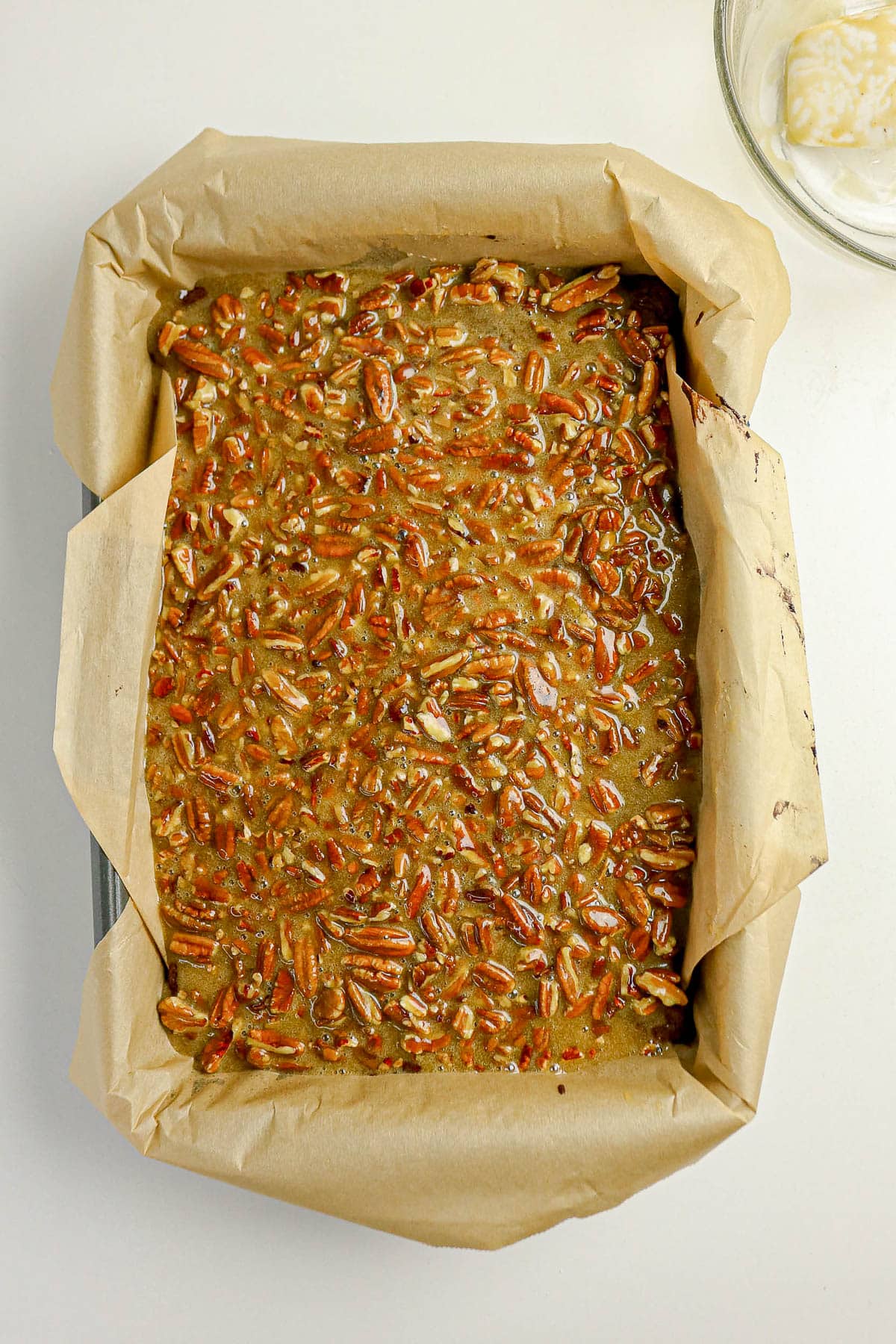 A baking pan lined with parchment paper, filled with unbaked pecan-topped batter on a white surface.