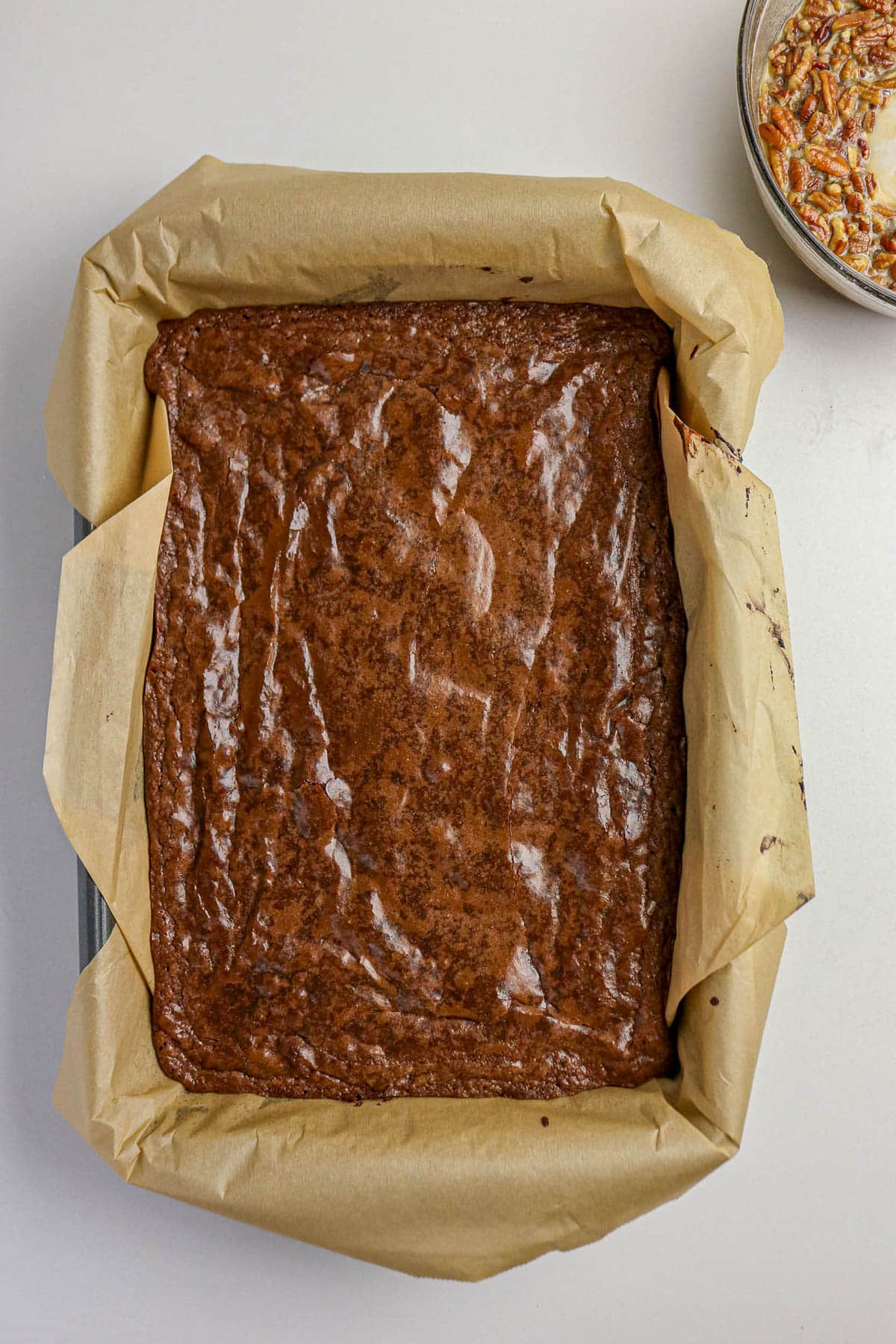 A pan of baked brownies lined with parchment paper, with a bowl of ingredients nearby on a white surface.